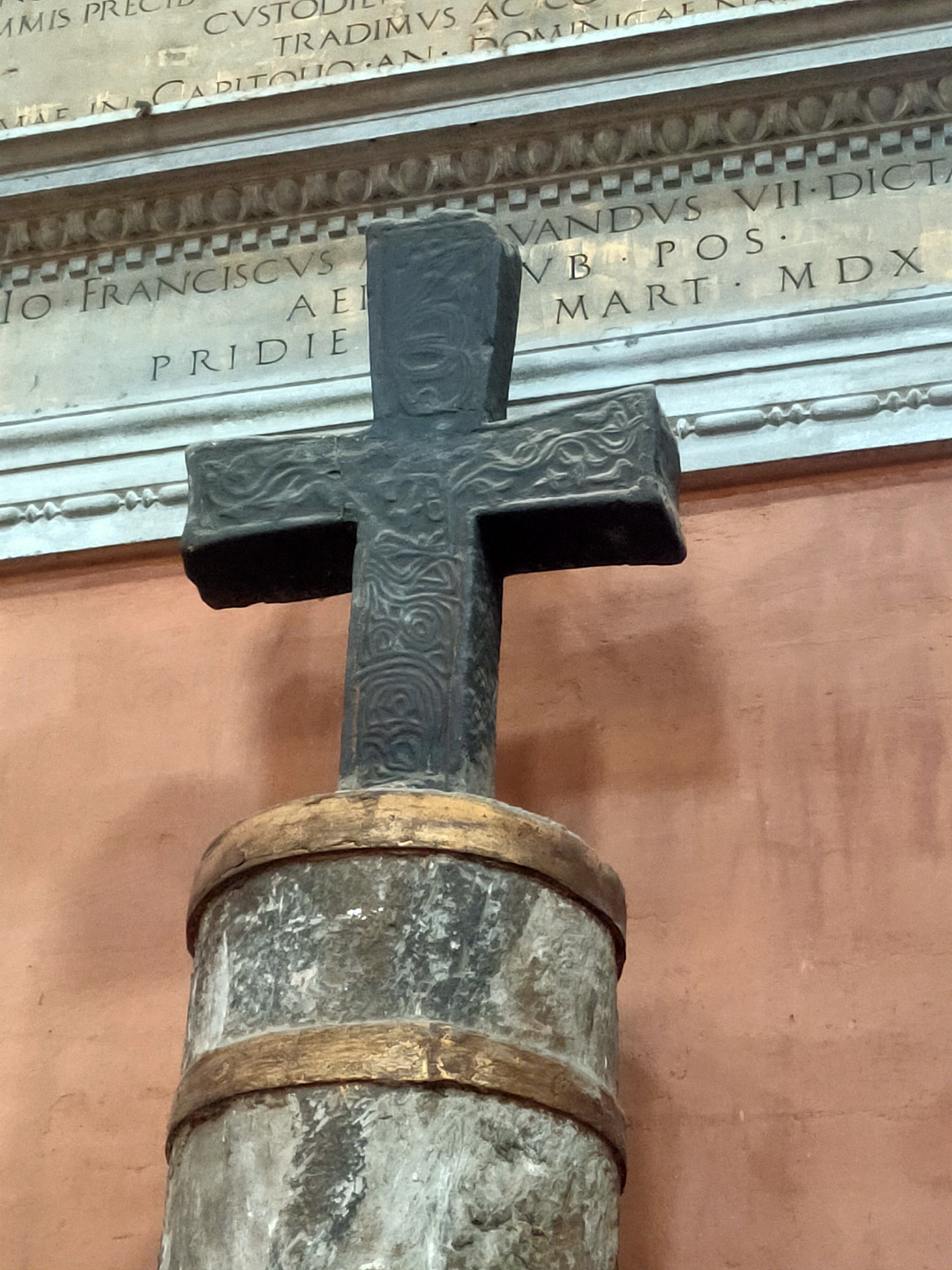 Croce scura decorata sopra una colonna, davanti a iscrizioni sulla parete