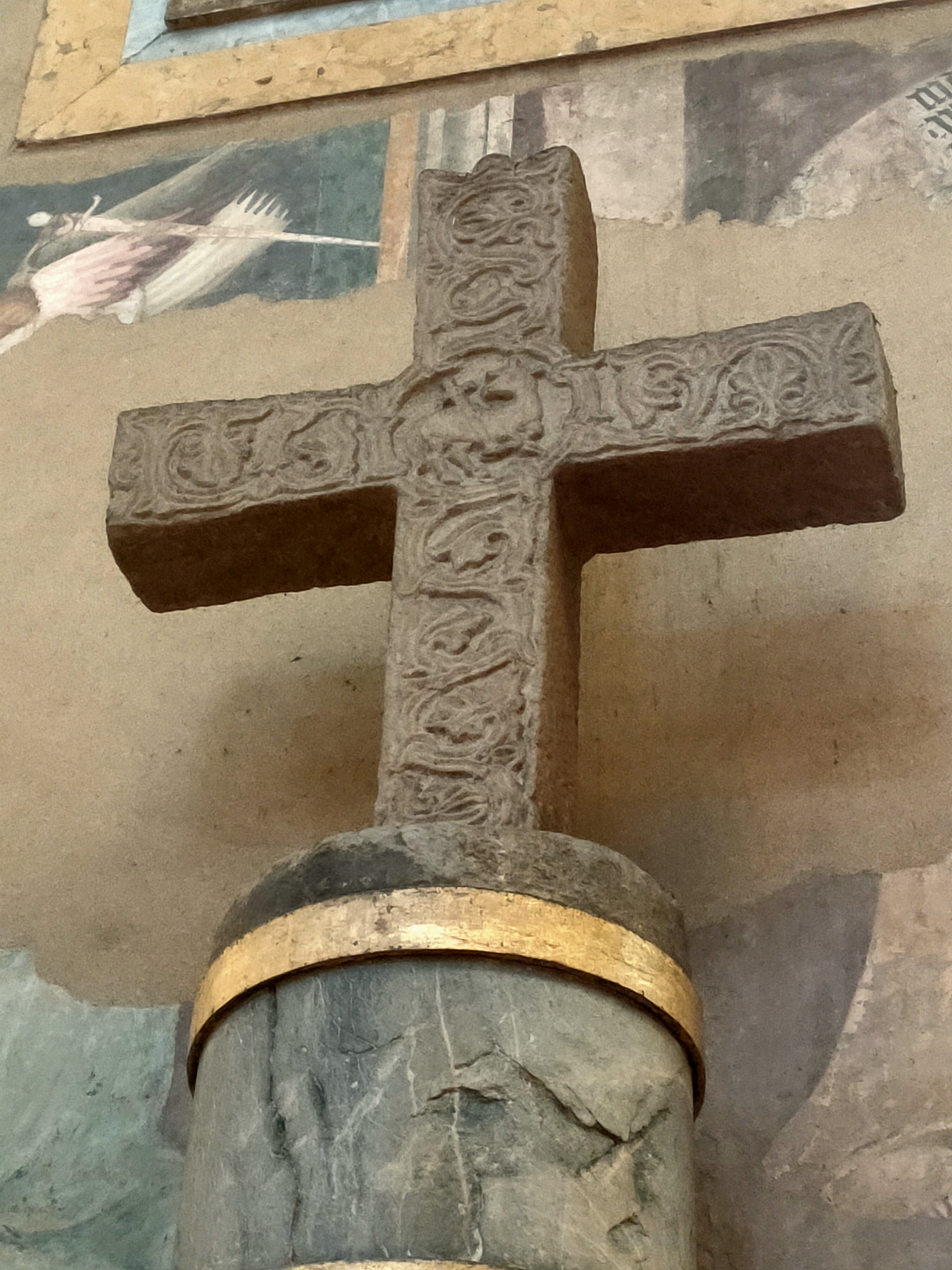 Croce in pietra decorata sopra una colonna scura davanti a un affresco